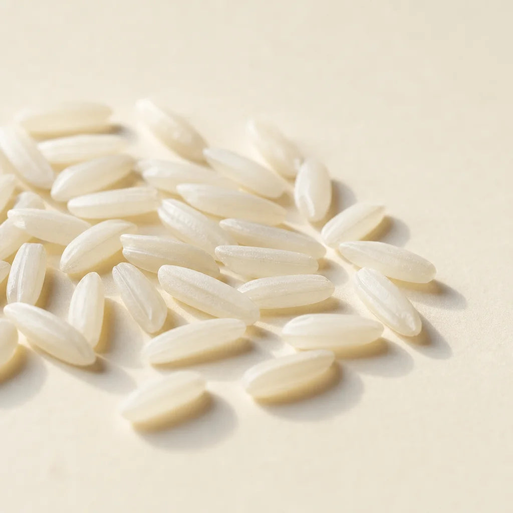 Clean editorial macro photography, square format. A cluster of raw white rice grains scattered light