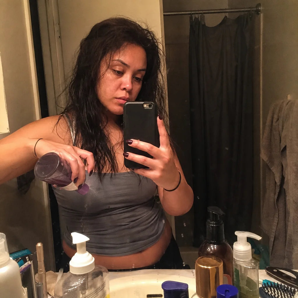 Square format, raw smartphone photograph. A bathroom mirror selfie of the same woman, now reapplying